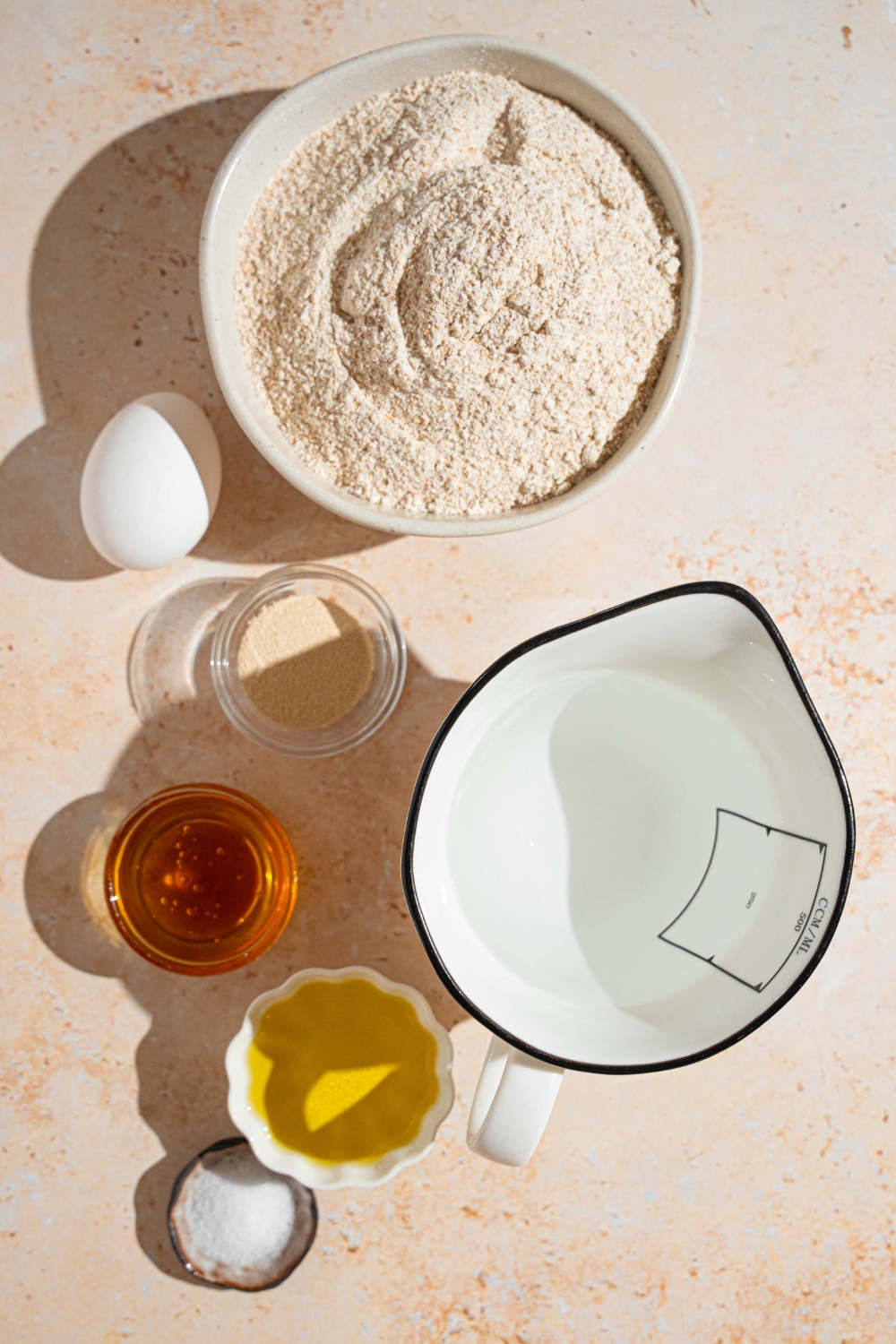 An overhead shot of several bowls in various sizes containing ingredients to make Ezekiel bread including Ezekiel mix, olive oil, honey, egg, salt, instant yeast, and water.