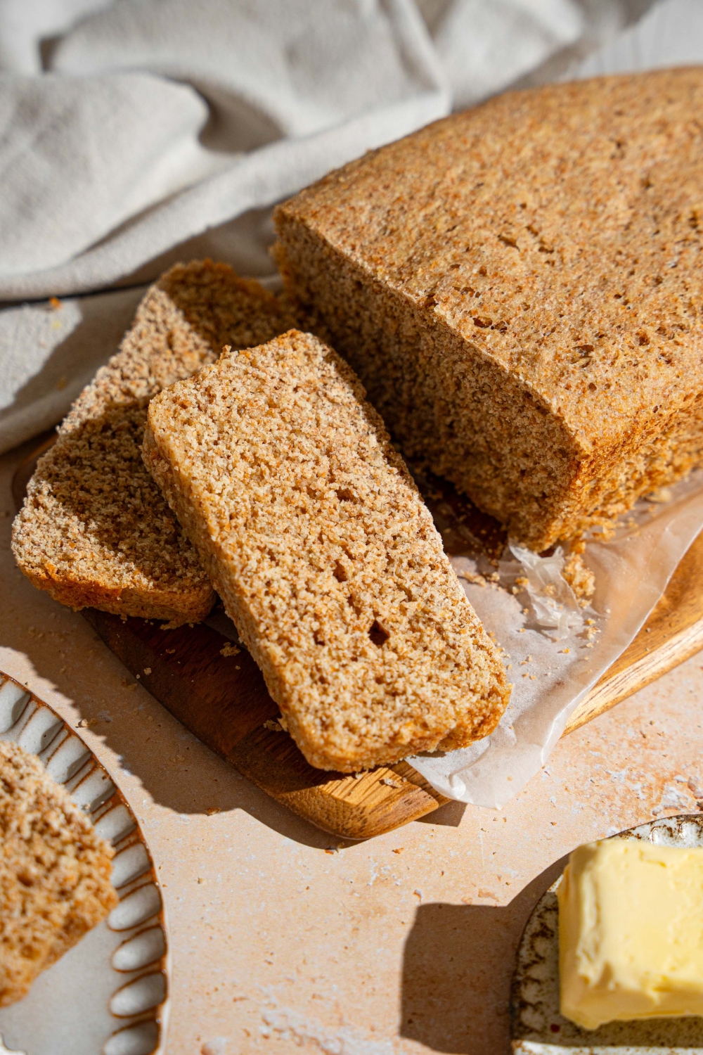 A loaf of Ezekiel bread on a wooden board lined with parchment paper. Two slices are sliced from the loaf. The board is on a tan counter with a white cloth napkin.