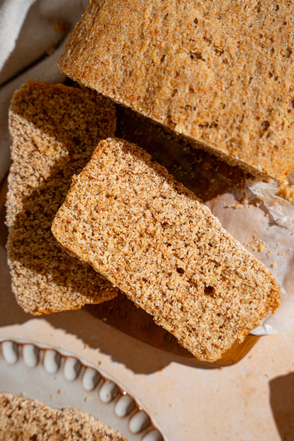 A loaf of Ezekiel bread on a wooden board lined with parchment paper. Two slices are sliced from the loaf. The board is on a tan counter with a white cloth napkin.