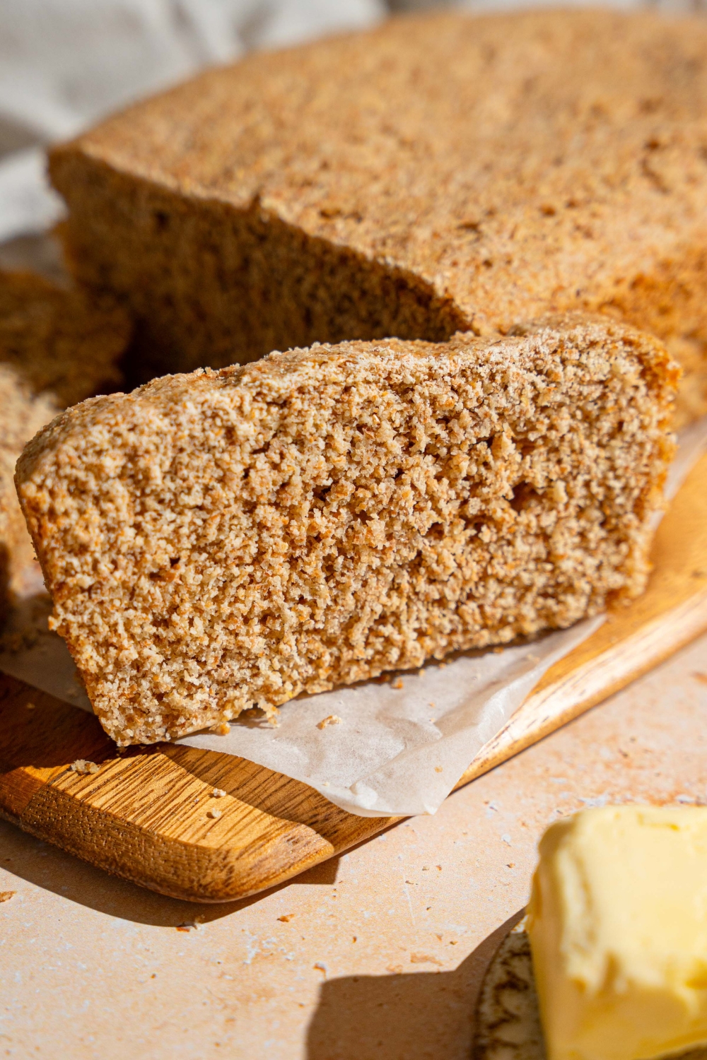 A loaf of Ezekiel bread on a wooden board lined with parchment paper. A slices is sliced from the loaf. The board is on a tan counter with a white cloth napkin and plate of butter.
