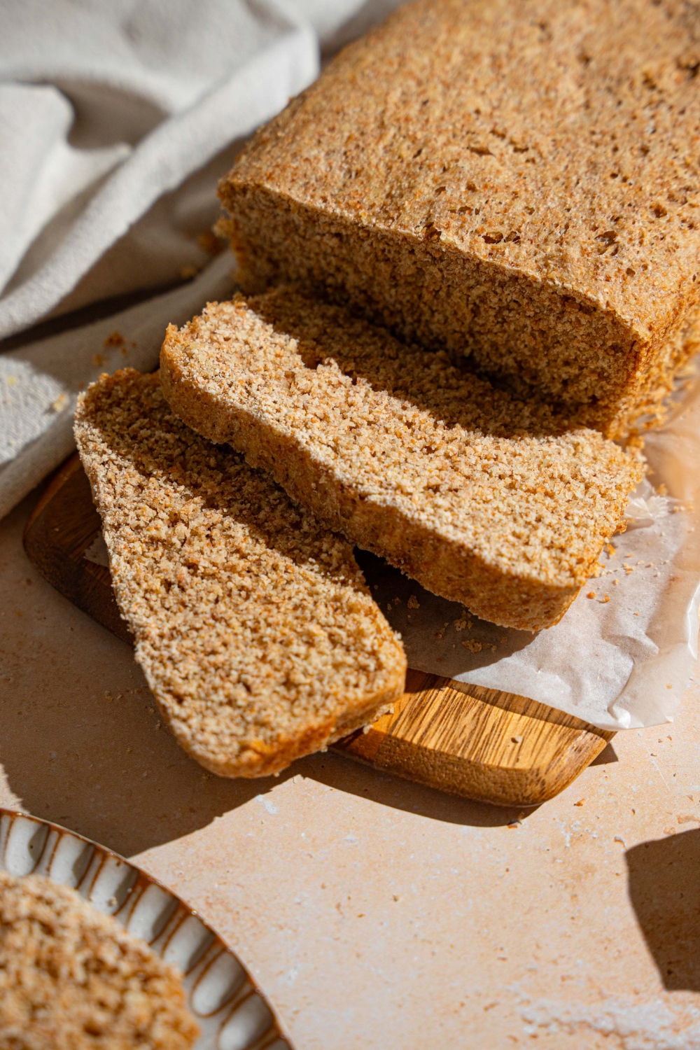 A loaf of Ezekiel bread on a wooden board lined with parchment paper. Two slices are sliced from the loaf. The board is on a tan counter with a white cloth napkin.