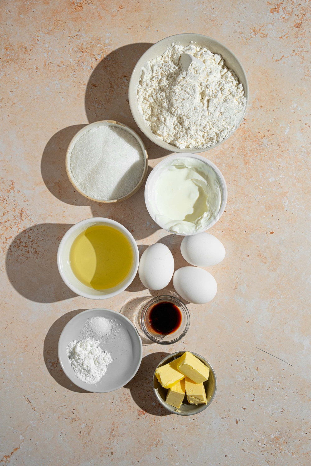 An overhead shot of several bowls in various sizes containing ingredients to make French butter cake including flour, yogurt, eggs, vanilla extract, sugar, butter, and baking powder.