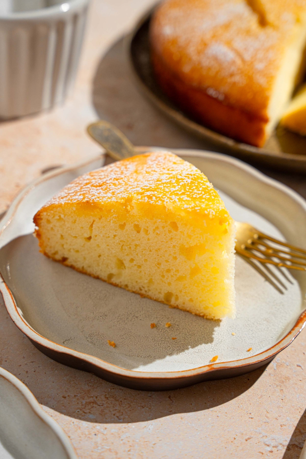 A white plate with a slice of French butter cake dusted with powdered sugar. There is a fork on the plate. The plate is on a tan counter with a whole cake.
