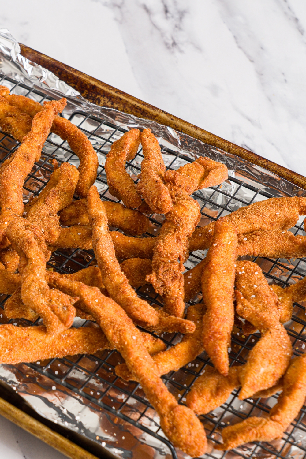A baking sheet lined with foil and with a rack with fried chicken fries seasoned with salt. The sheet is on a marble counter.