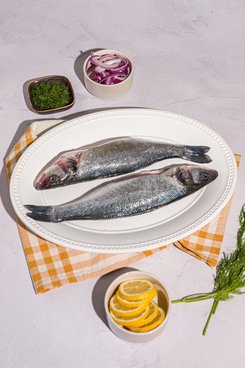 A white plate with two whole branzinos on a white counter with small bowls of lemon, herbs, and sliced red onion.