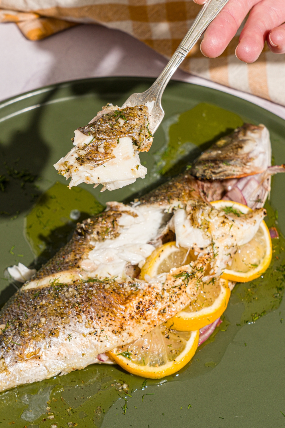 A green plate with greek whole roasted branzino stuffed with sliced lemon. There is a fork with a bite of branzino held over the plate. The plate is on a white counter with a yellow checkered napkin.