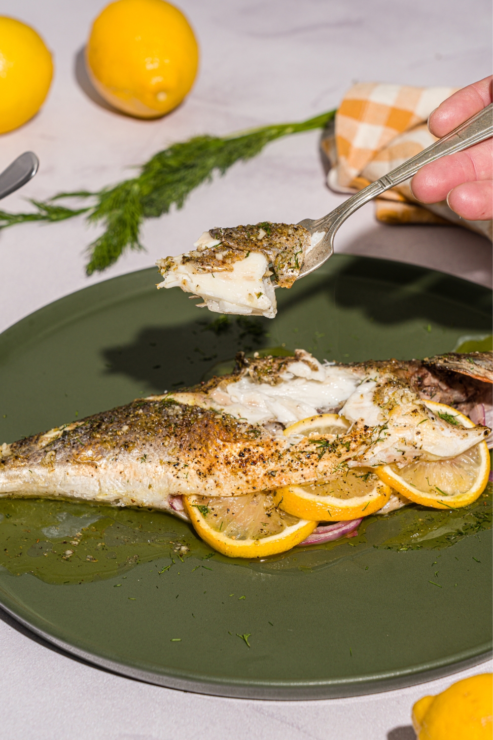 A green plate with greek whole roasted branzino stuffed with sliced lemon. There is a fork with a bite of branzino held over the plate. The plate is on a white counter with a yellow checkered napkin.