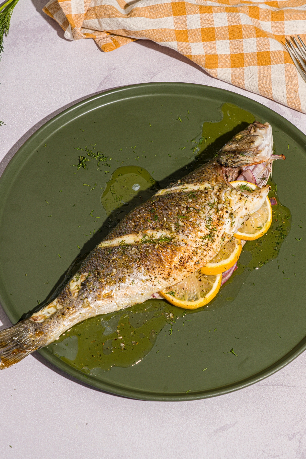 A green plate with greek whole roasted branzino stuffed with sliced lemon. The plate is on a white counter with a yellow checkered napkin.