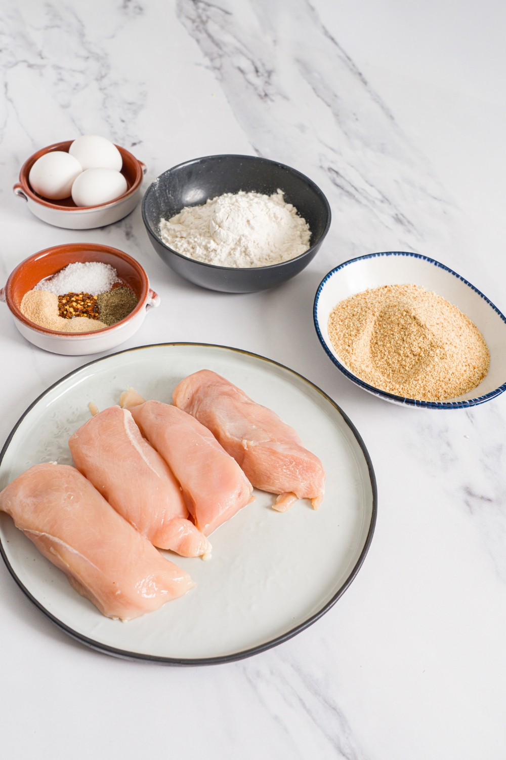 Several bowls in various sizes containing ingredients to make homemade chicken fries including chicken breasts, flour, breadcrumbs, eggs, and seasonings.