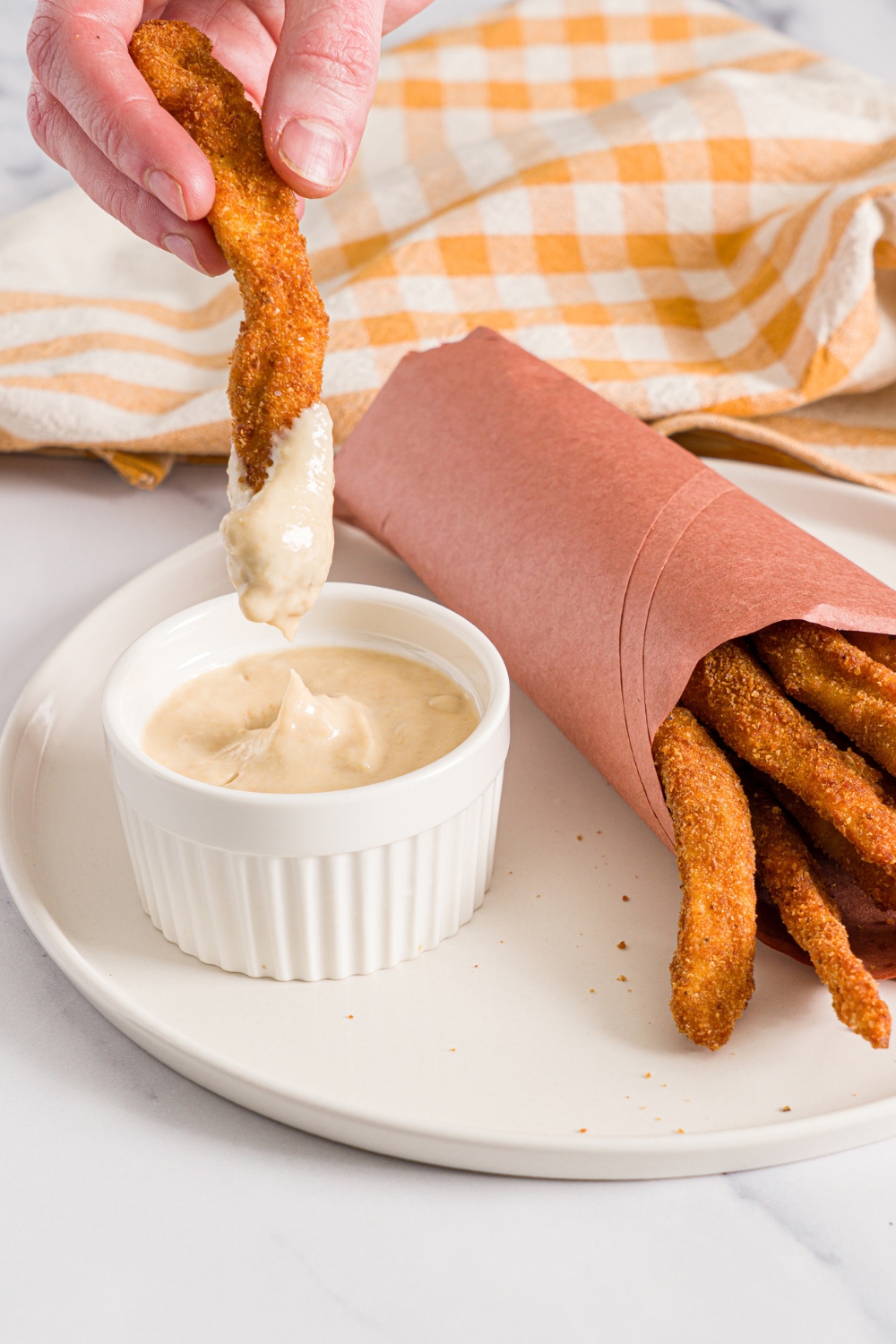 A white plate with fried homemade chicken fries wrapped in contact paper with a small ramekin of garlic aioli. A hand is dipping a chicken fry into the aioli.The plate is on a marble counter with a yellow checkered napkin.