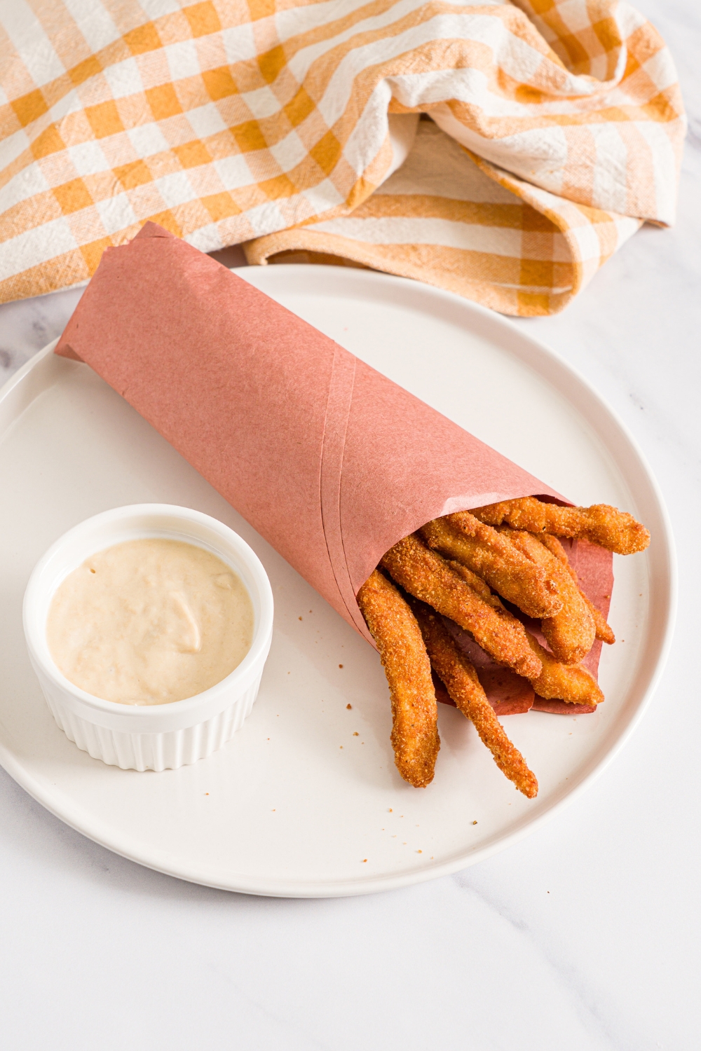A white plate with fried homemade chicken fries wrapped in contact paper with a small ramekin of garlic aioli. The plate is on a marble counter with a yellow checkered napkin.