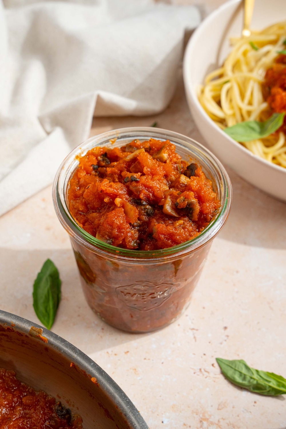 A jar of homemade spaghetti sauce with fresh tomatoes. The jar is on a tan counter with a bowl of spaghetti and fresh basil with a white cloth napkin.