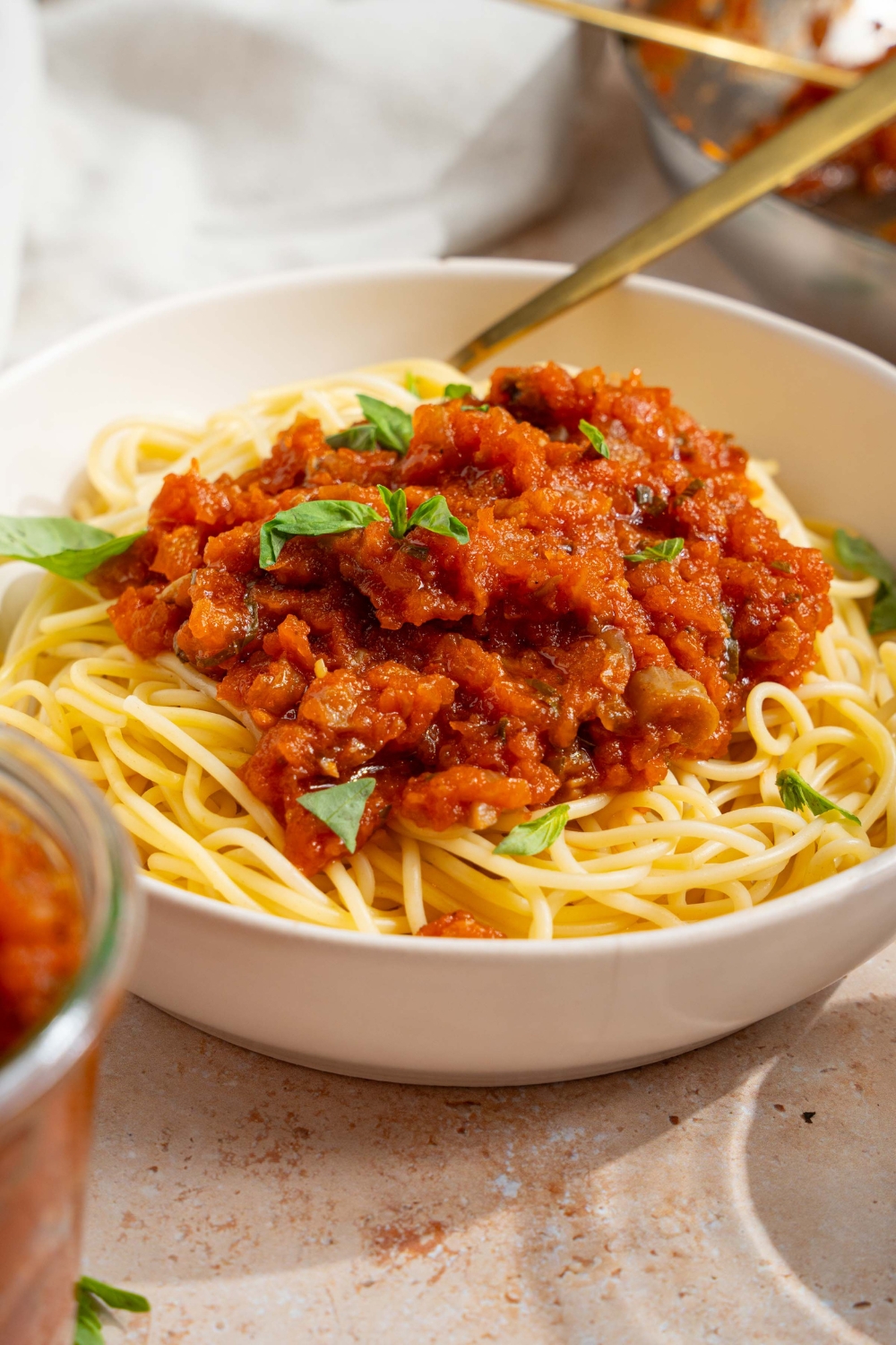 A bowl of spaghetti with homemade spaghetti sauce with fresh tomatoes. The bowl is garnished with fresh basil. There is a fork in the bowl. The bowl is on a tan counter with a white cloth napkin.