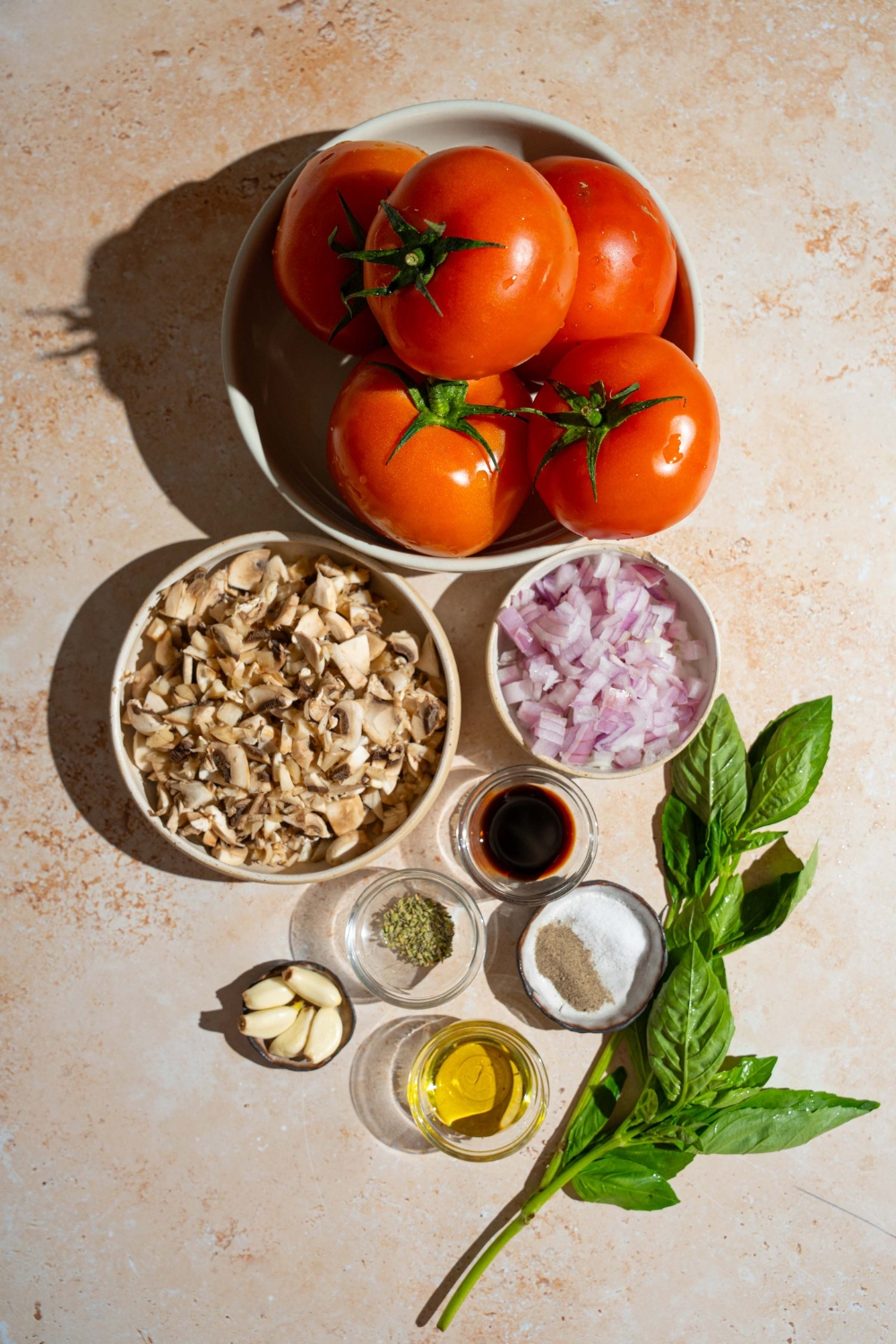 An overhead shot of several bowls in various sizes containing ingredients to make homemade spaghetti sauce with fresh tomatoes including tomatoes, onion, garlic, mushrooms, soy sauce, oil, and seasonings.