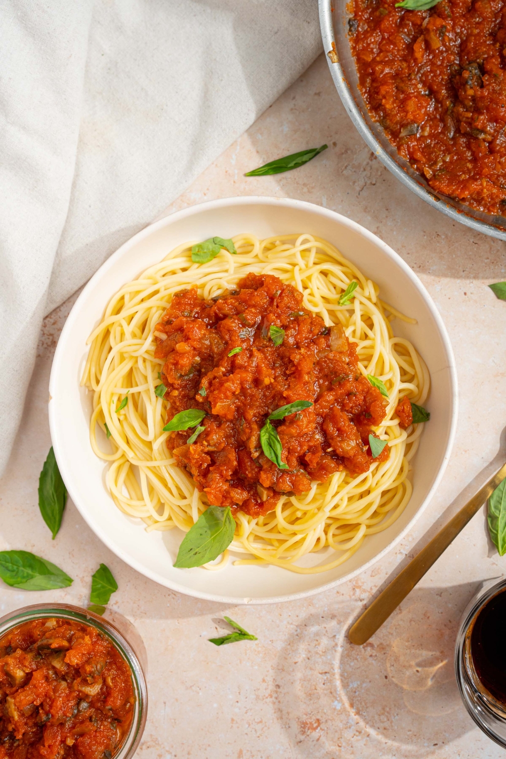 A bowl of spaghetti with homemade spaghetti sauce with fresh tomatoes. The bowl is garnished with fresh basil. The bowl is on a tan counter with a white cloth napkin and skillet of sauce.