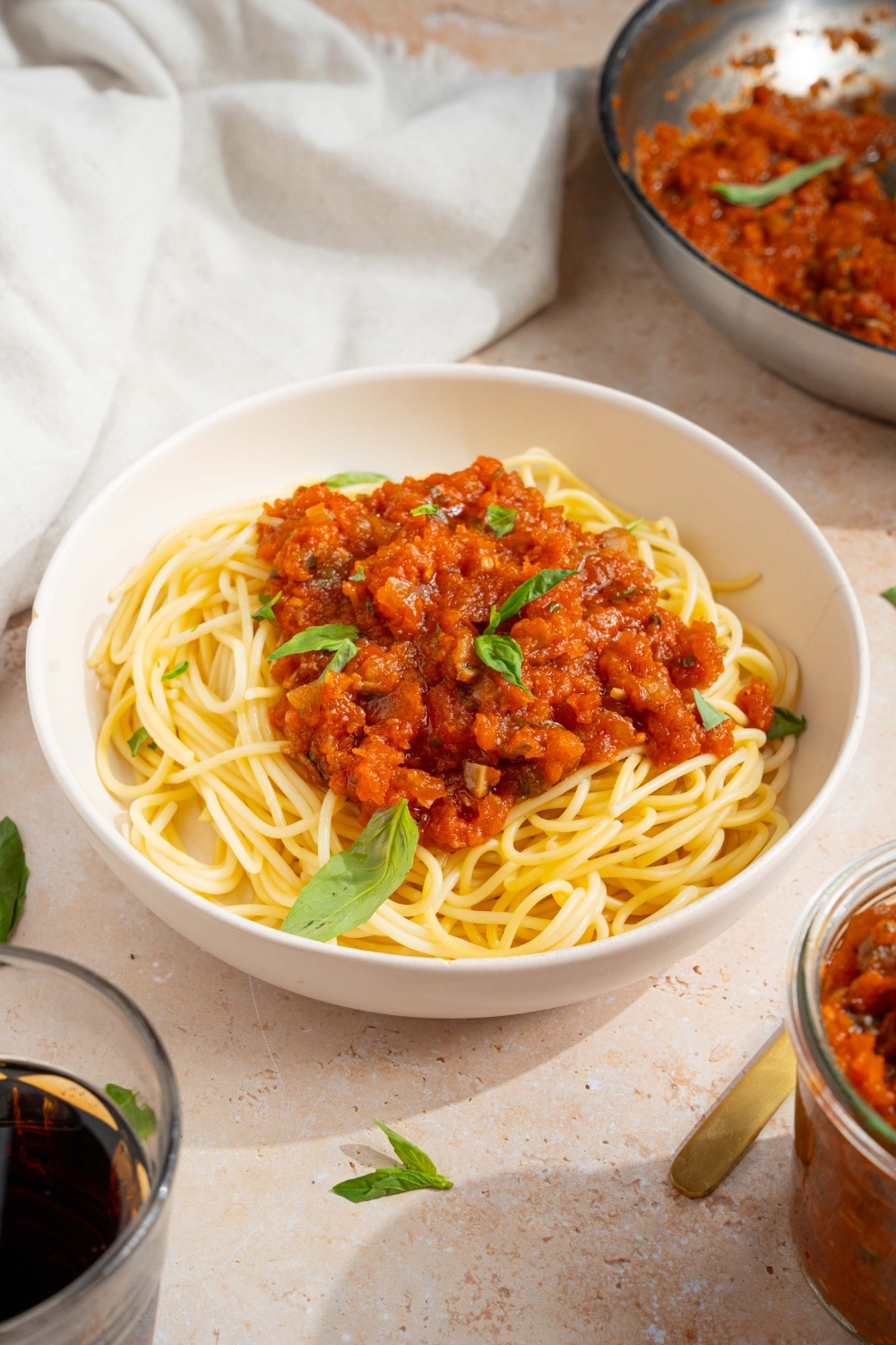`A bowl of spaghetti with homemade spaghetti sauce with fresh tomatoes. The bowl is garnished with fresh basil. The bowl is on a tan counter with a white cloth napkin and skillet of sauce.