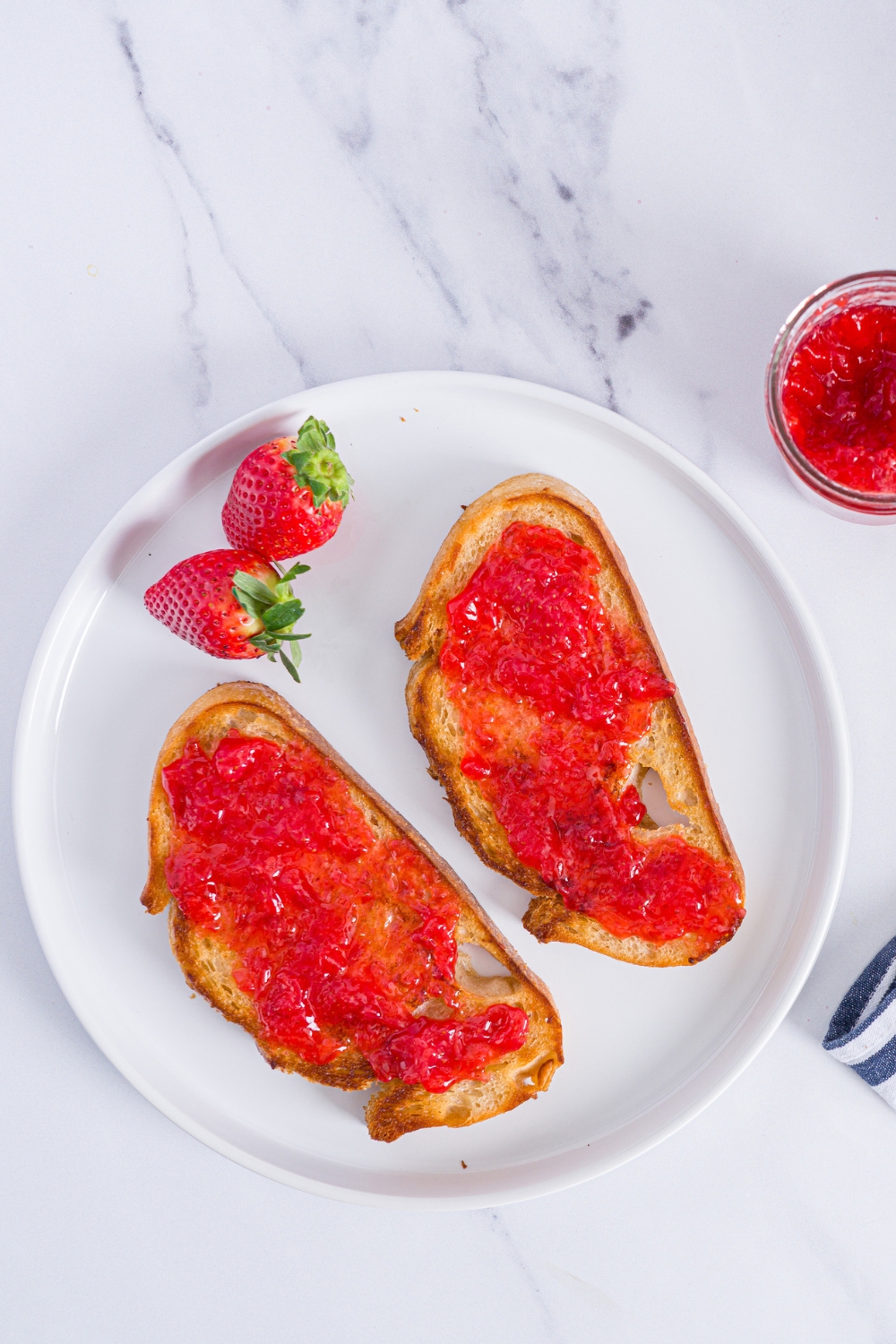 A white plate with two slices of toast with strawberry jam and strawberries. The plate is on a marble counter with a jar of jam.