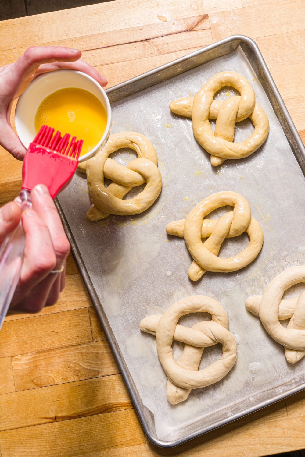 A baking sheet lined with parchment paper with unbaked Auntie Anne's pretzels. A hand is brushing egg wash with a brush on the pretzels.