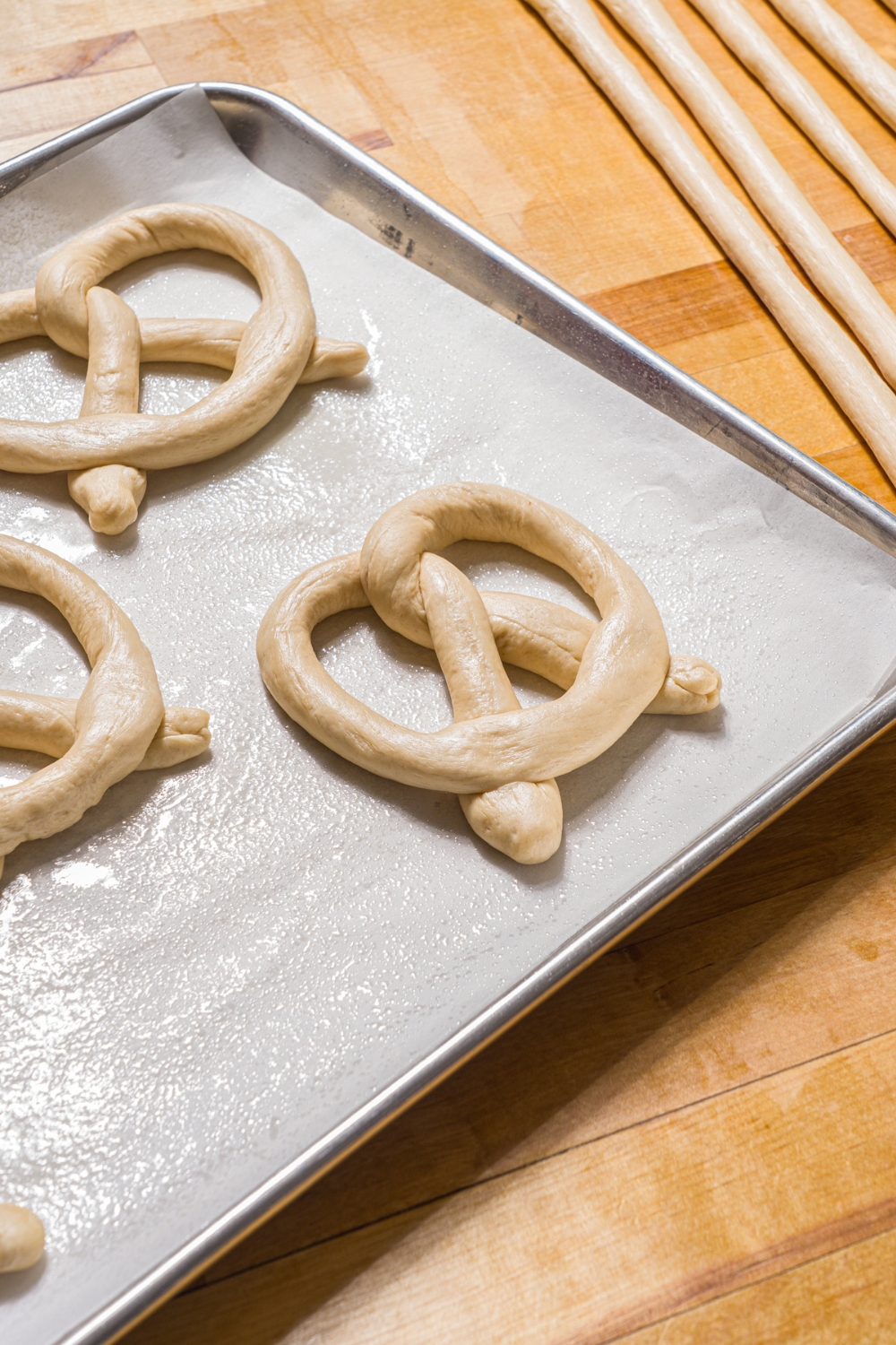 A baking sheet lined with parchment paper with unbaked Auntie Anne's pretzels. The baking sheet is on a wood counter with strips of pretzel dough.