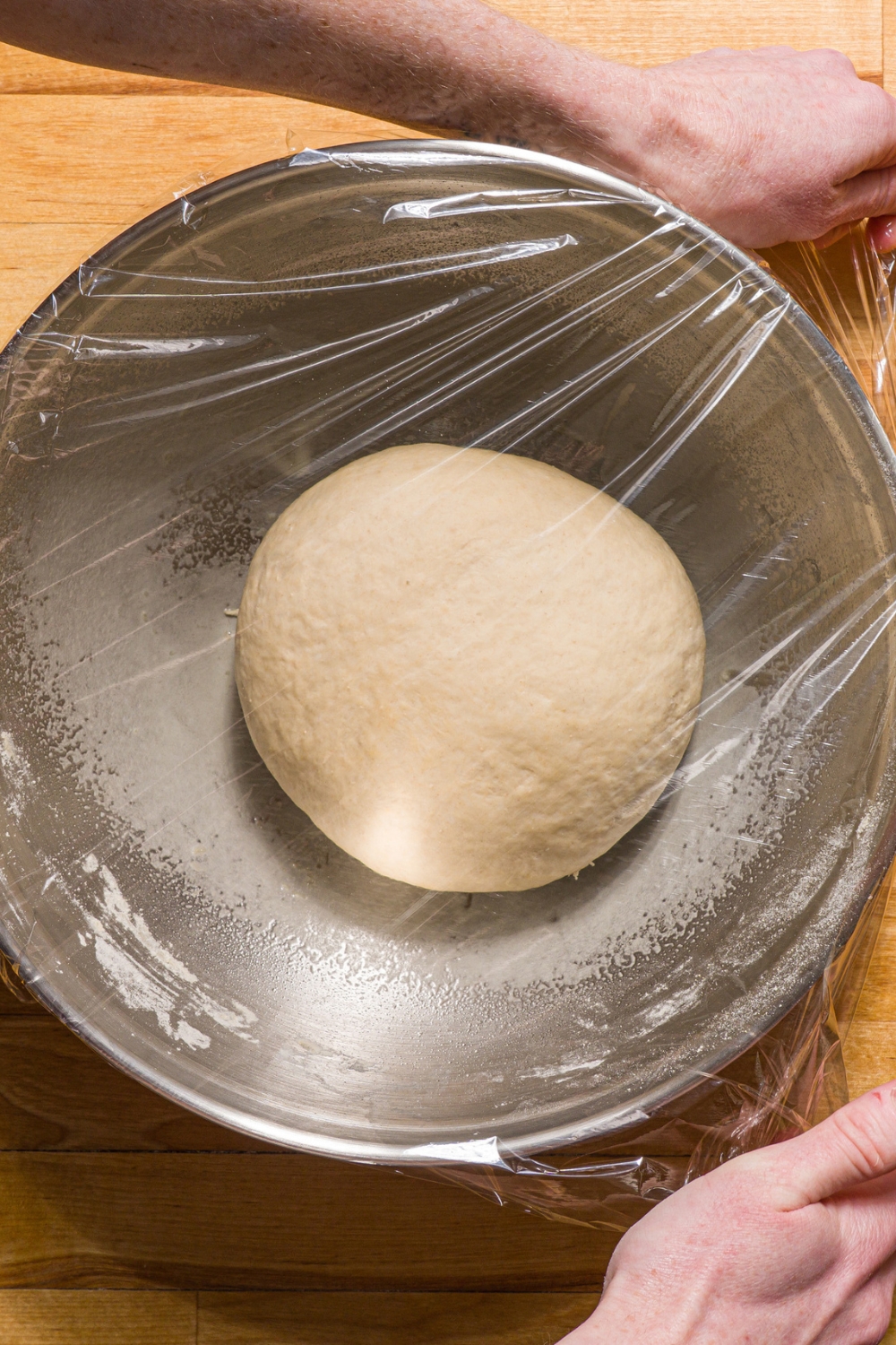 A large bowl with a ball of kneaded pretzel dough. The bowl is being covered with saran wrap.