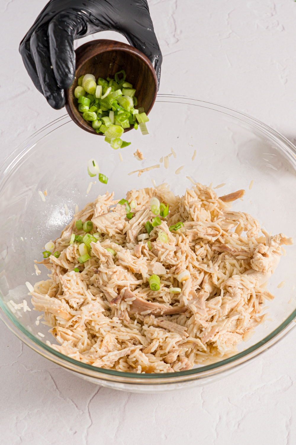 A glass bowl with shredded chicken tossed with rice. A small bowl of sliced scallions are being added to the chicken. The bowl is on a white counter.