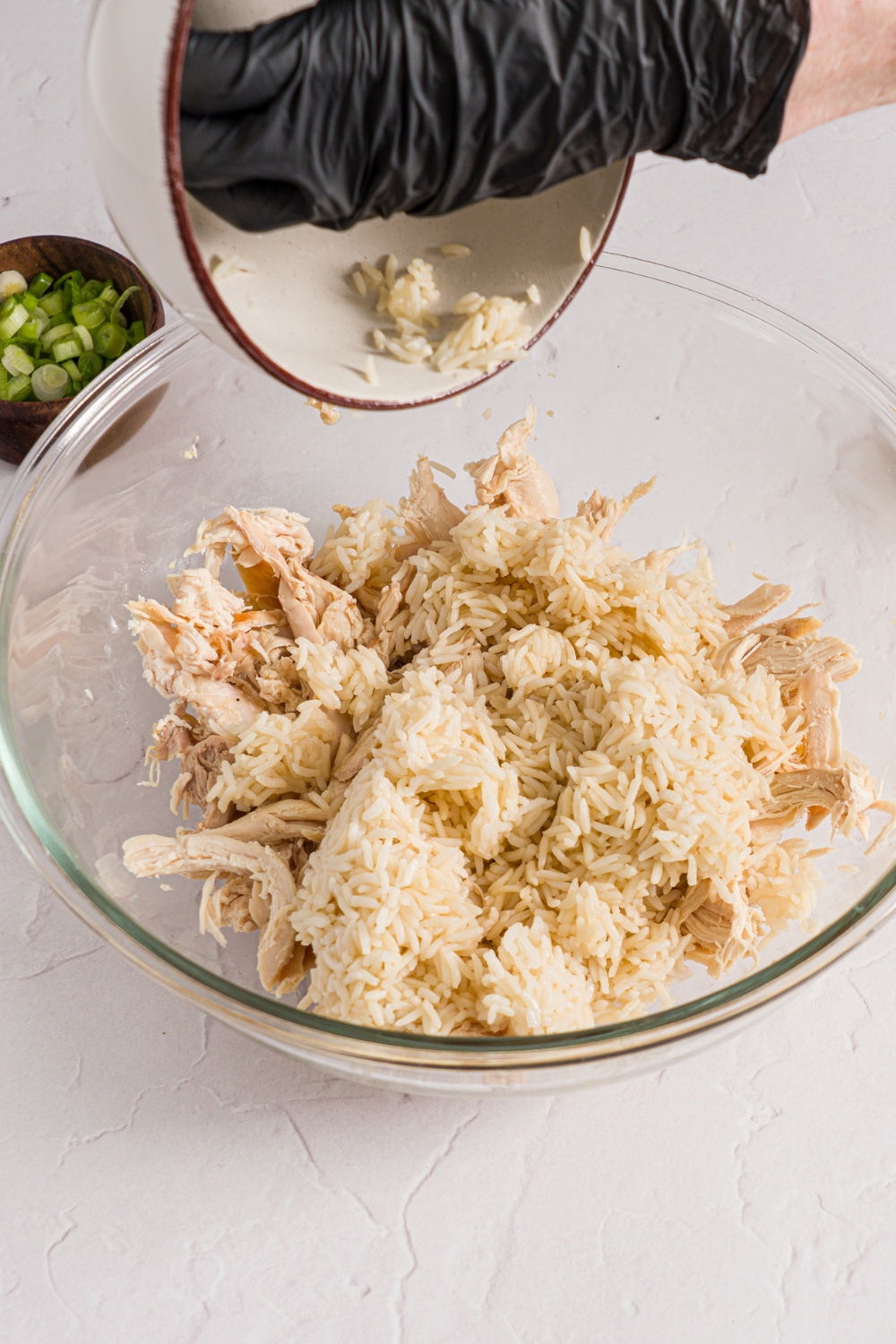 A glass bowl with shredded chicken with a small bowl of white rice being added to the bowl. The bowl is on a white counter with a small bowl of sliced scallions.