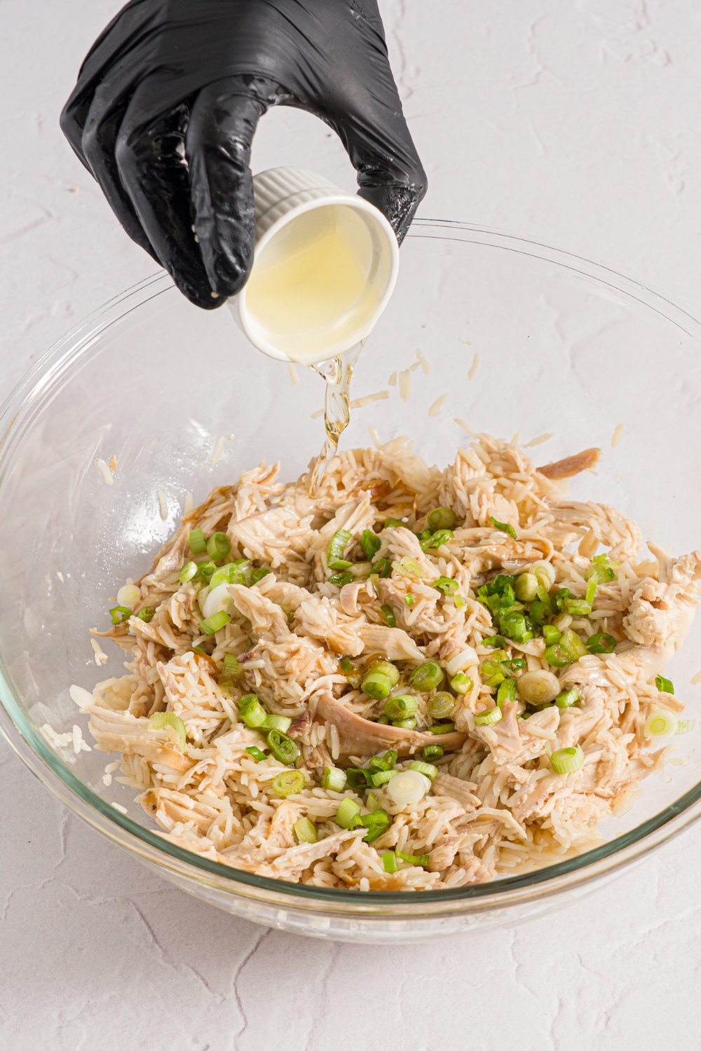 A glass bowl with a chicken, scallion, and rice mixture with a small bowl of oil being added to the mixture. The bowl is on a white counter.