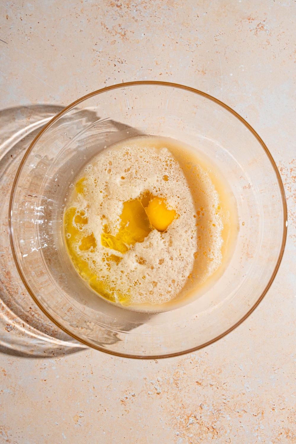 A glass bowl with eggs and oil mixed with a yeast mixture. The bowl is on a tan counter.