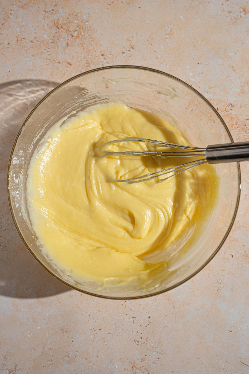 A glass bowl with a whisk mixing French butter cake batter. The bowl is on a tan counter.