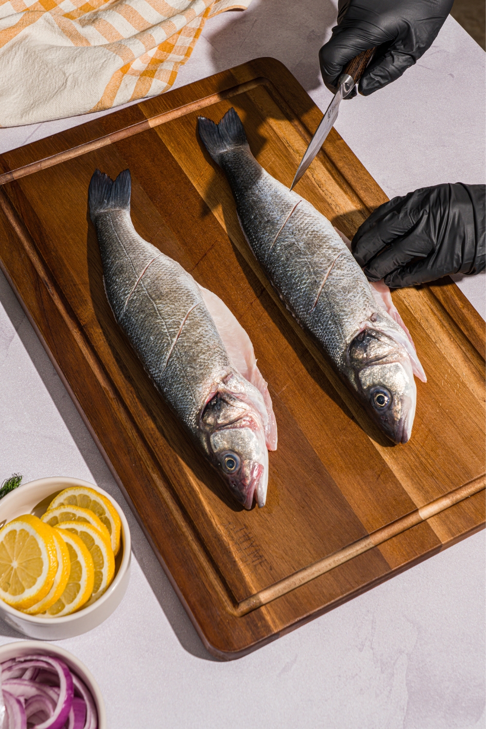 A wooden board with two whole branzinos. The branzinos are being slit with a knife. The board is on a white counter with a bowl of sliced lemons and yellow checkered napkin.