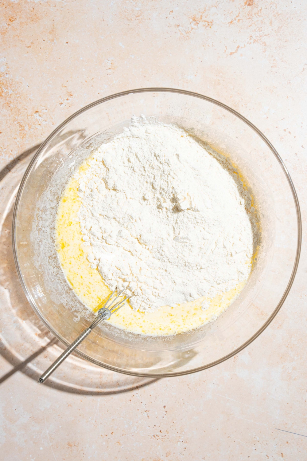 A bowl with wet and dry ingredients to make IHOP New York cheesecake pancakes. There is a whisk in the bowl. The bowl is on a tan counter.