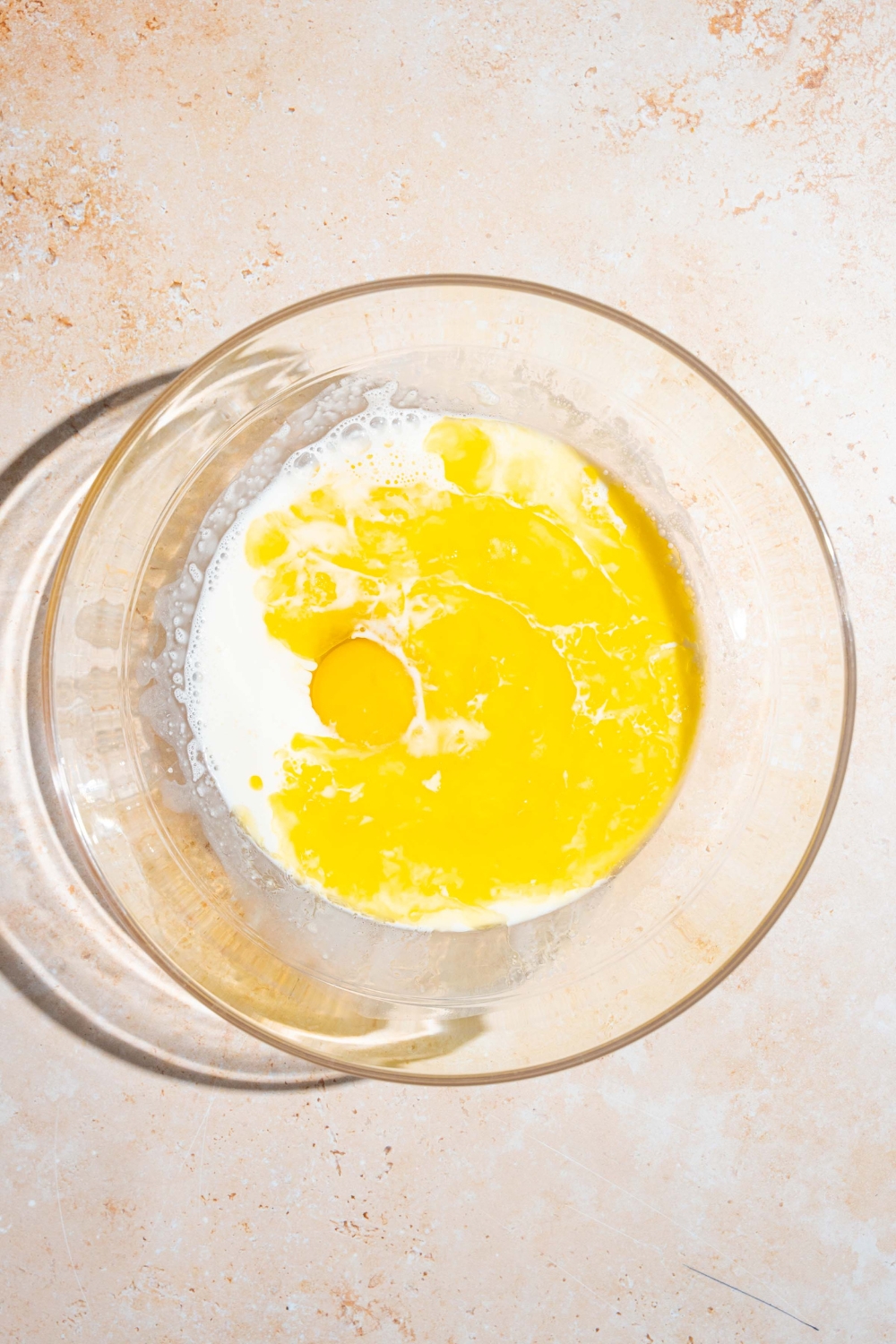 A glass bowl with wet ingredients to make IHOP New York cheesecake pancake batter including egg, milk, and butter. The bowl is on a tan counter.
