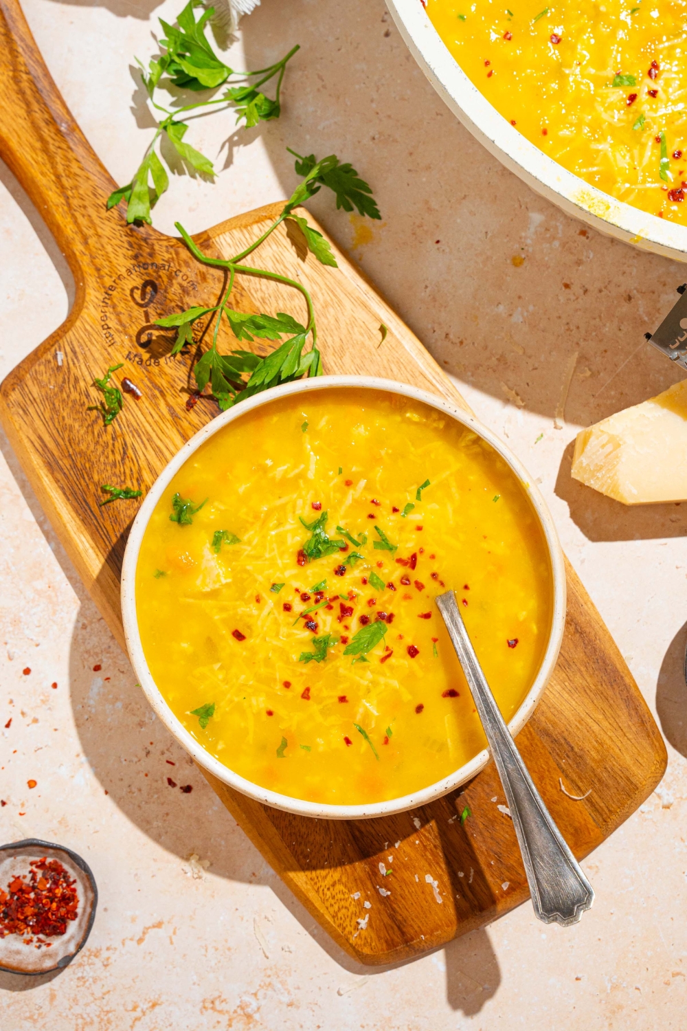 A white bowl of Italian penicillin soup garnished with fresh parsley and crushed red pepper. There is a spoon in the bowl. The bowl is on a wooden board on a white counter with a pot of soup and fresh parsley.