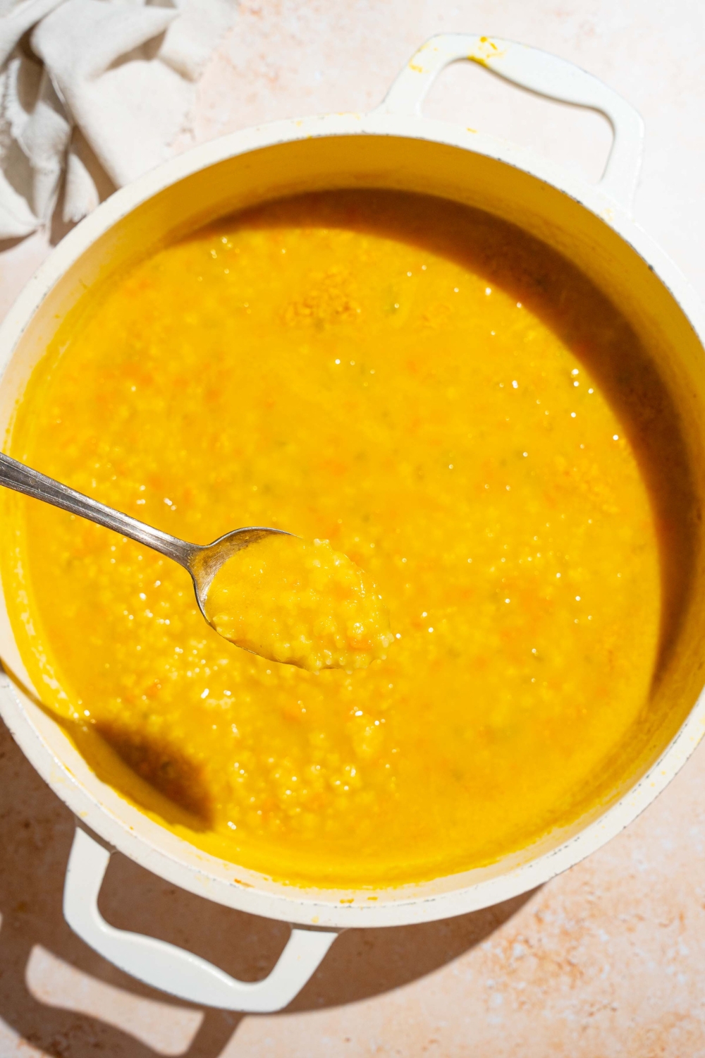 A stock pot with Italian penicillin soup with pastina cooking in the broth. There is a spoon in the pot with a bite of soup. The pot is on a tan counter with a white cloth napkin.