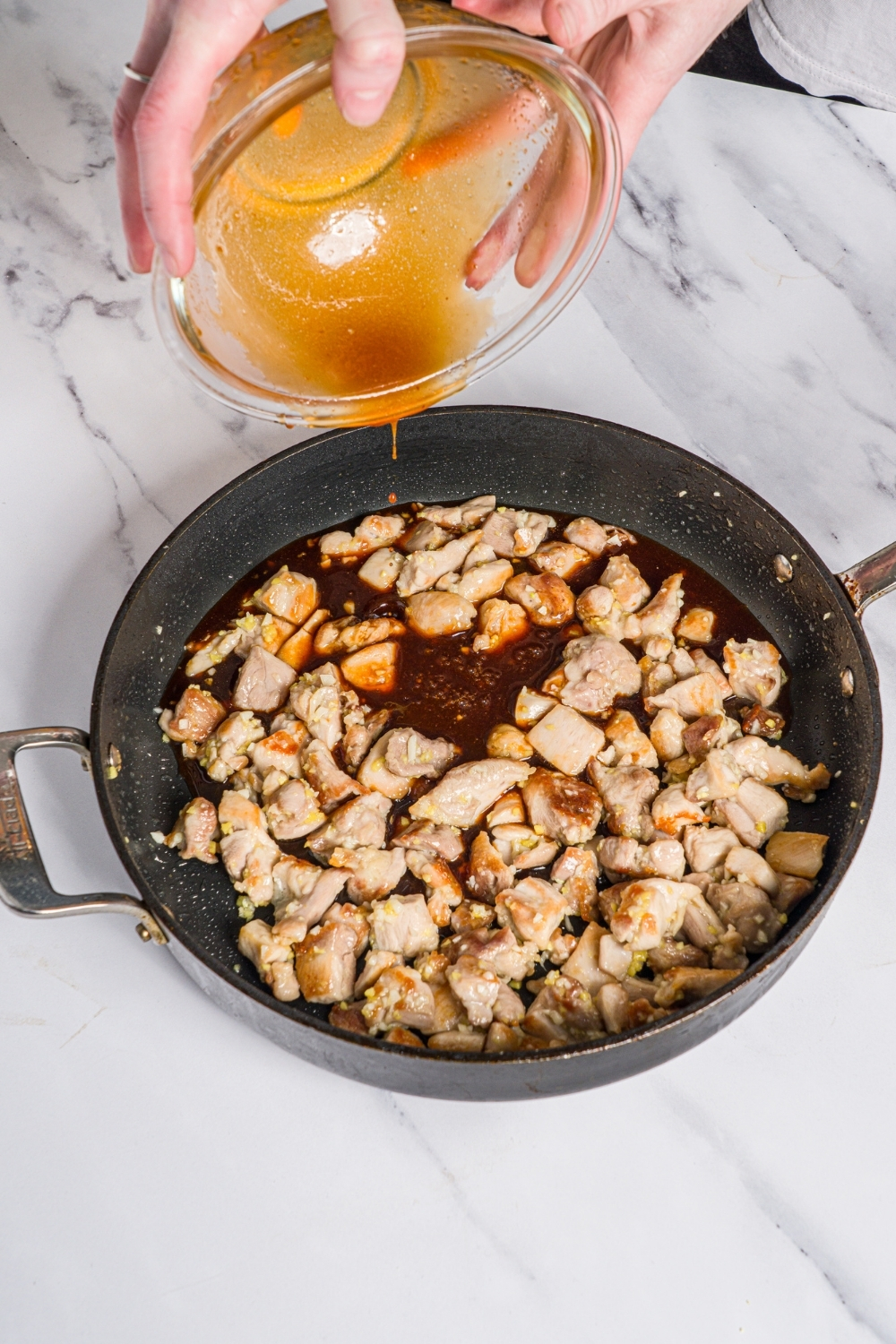 A skillet with cooked cubed chicken thighs with a bowl of Korean sticky chicken sauce being added to the skillet. The skillet is on a marble counter.