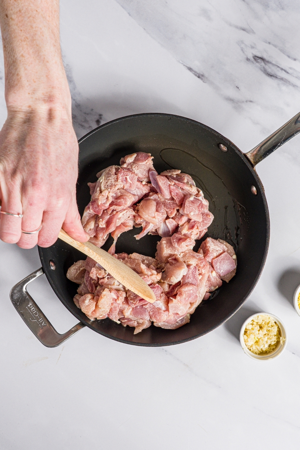 A skillet with uncooked cubed chicken thighs being broken up with a wooden spoon. The skillet is on a marble counter.