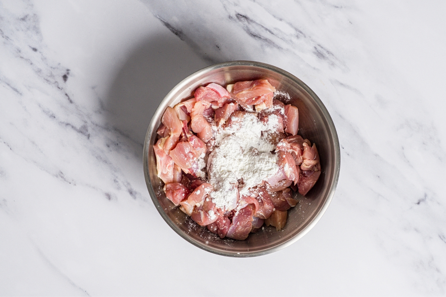 A bowl with uncooked cubed chicken thighs topped with corn starch. The bowl is on a marble counter.