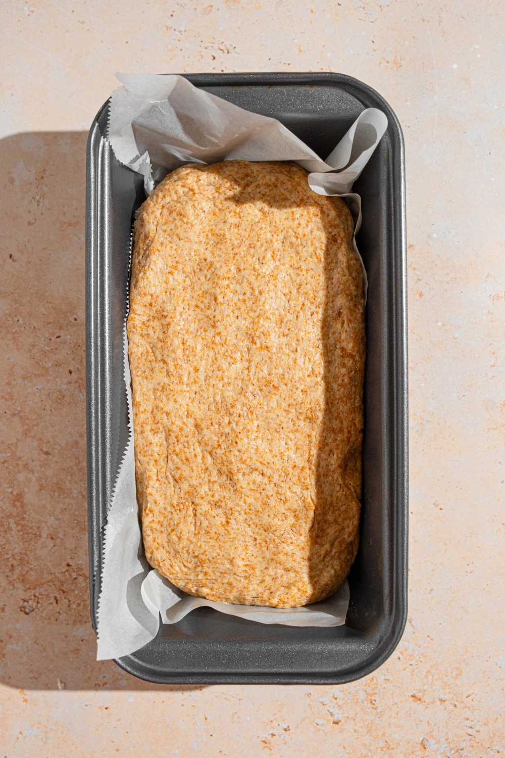 A loaf pan lined with parchment paper with Ezekiel bread dough. The pan is on a tan counter.