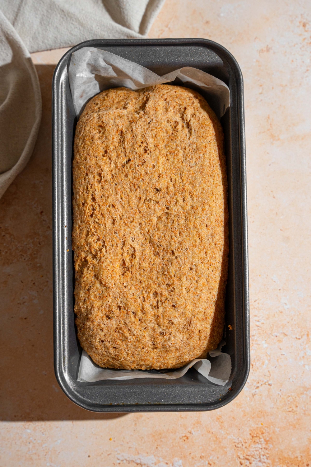 A loaf pan lined with parchment paper with baked Ezekiel bread. The pan is on a tan counter.