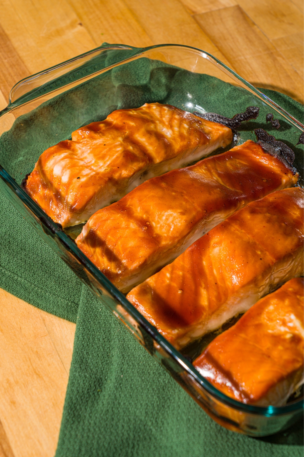 A glass baking dish with four baked teriyaki salmon fillets. The dish is on a green cloth napkin on a wood counter.