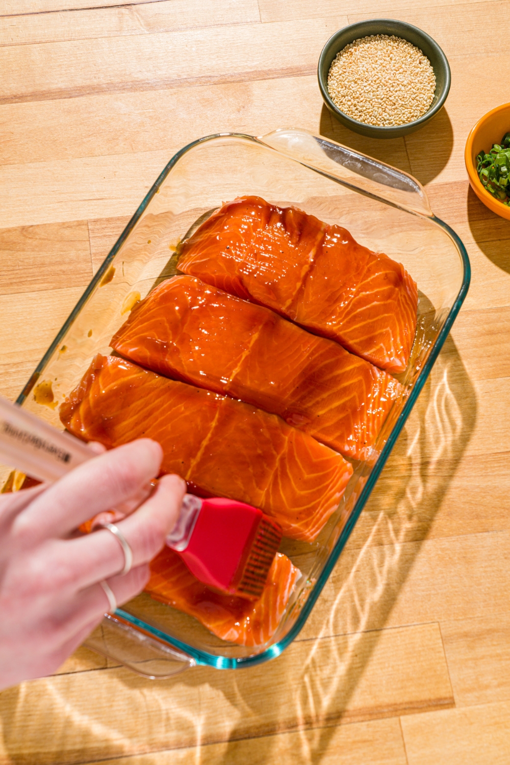 A glass baking dish with a brush brushing teriyaki sauce over uncooked salmon fillets. The dish is on a wood counter with small bowls of garnishes.