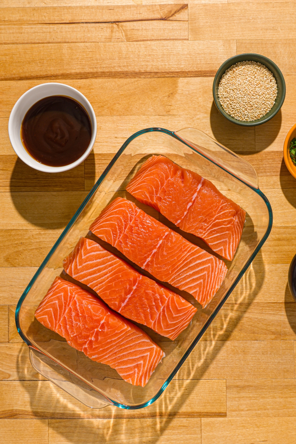 A baking dish with uncooked salmon fillets on a wood counter with a small bowl of teriyaki sauce and bowl of sesame seeds.