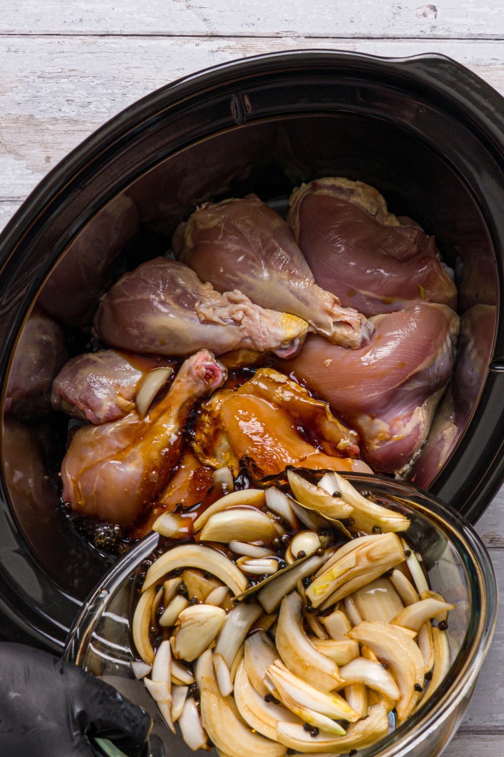 A crock pot with uncooked chicken with a bowl of onion and garlic sauce being poured over the chicken.
