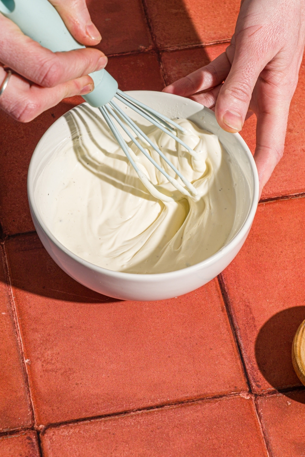 A white bowl with a hand whisking ingredients to make creamy lime dip. The bowl is on a red tiled counter.