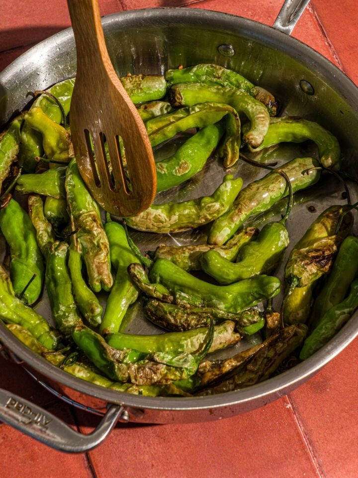 A skillet with shishito peppers blistering in oil. A wooden spoon is stirring the peppers.