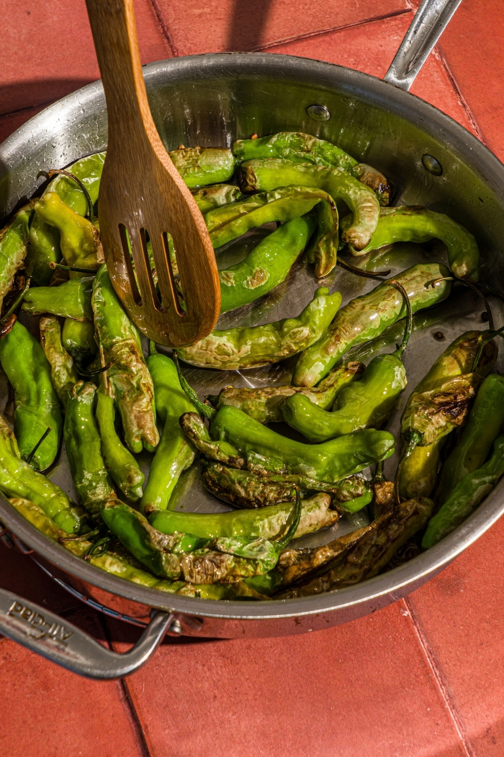 A skillet with shishito peppers blistering in oil. A wooden spoon is stirring the peppers.