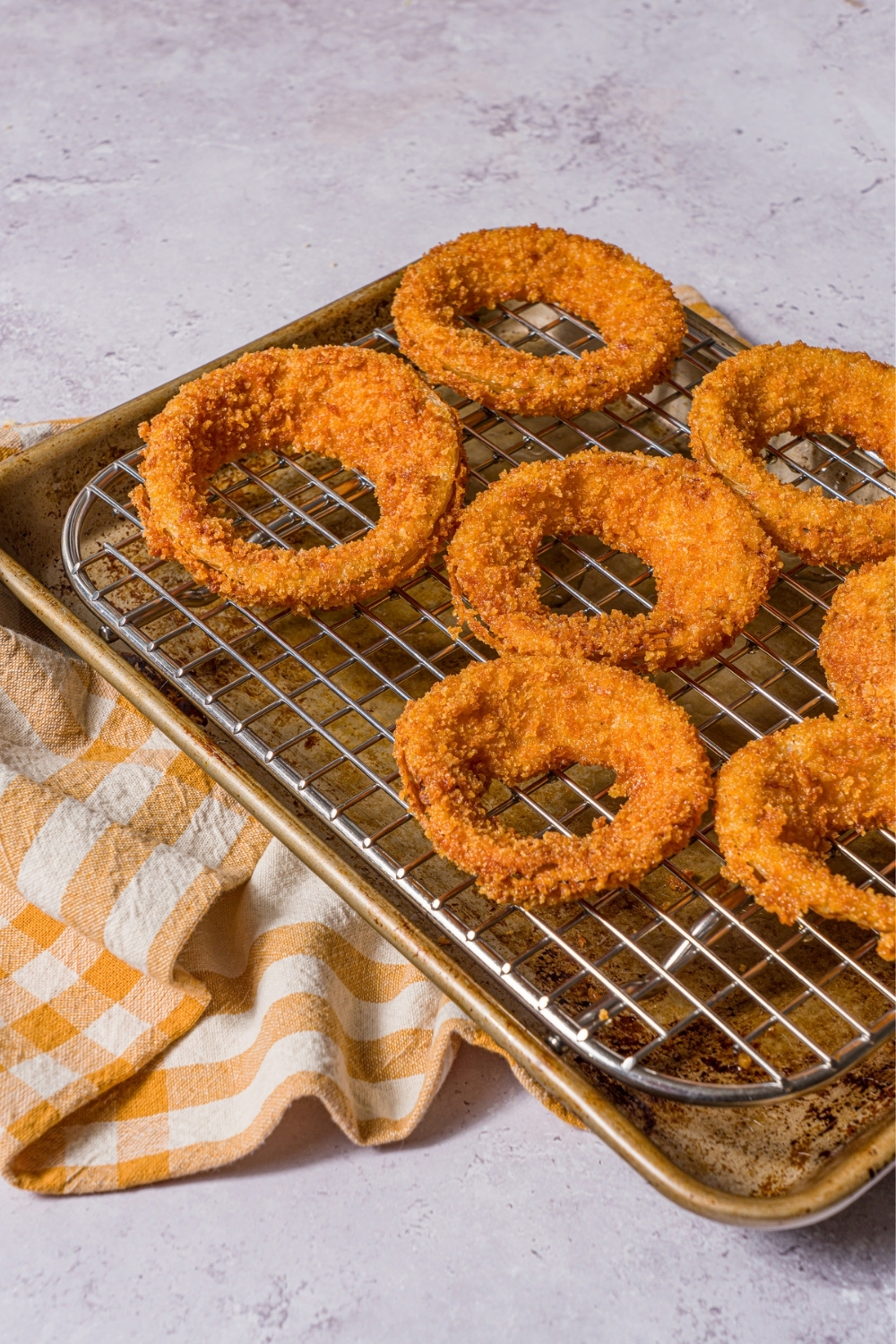 A baking sheet lined with a rack with fried onion rings. The sheet is on a stone counter with a yellow striped napkin.