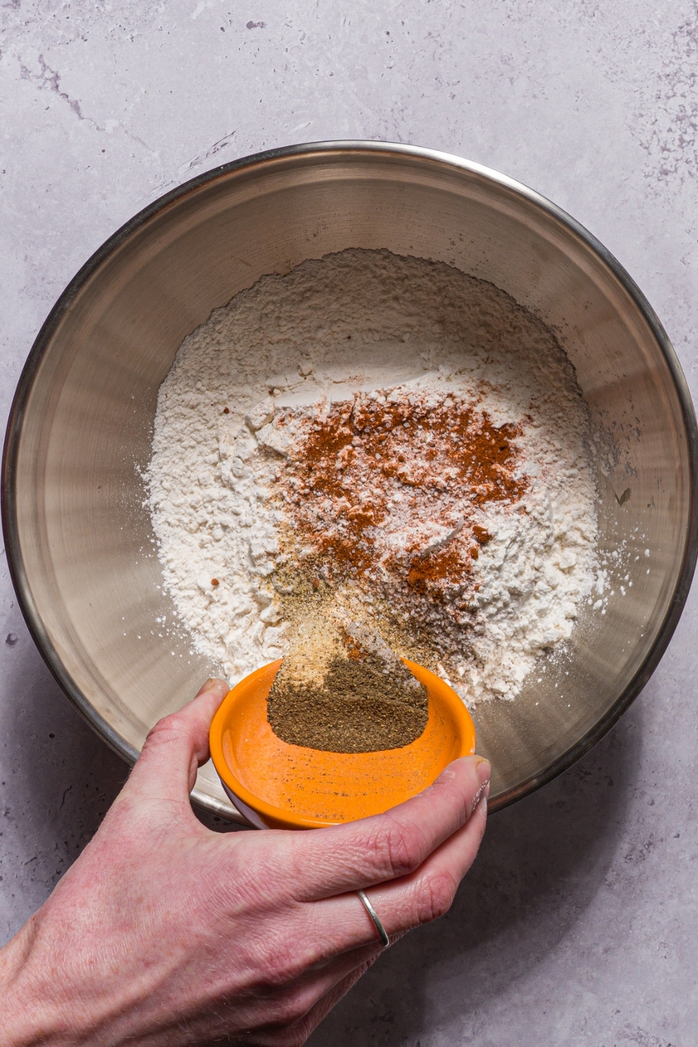 A bowl with ingredients to make a flour mixture for breading including flour, paprika, garlic powder, salt, and pepper. A hand is dumping seasonings into the bowl.