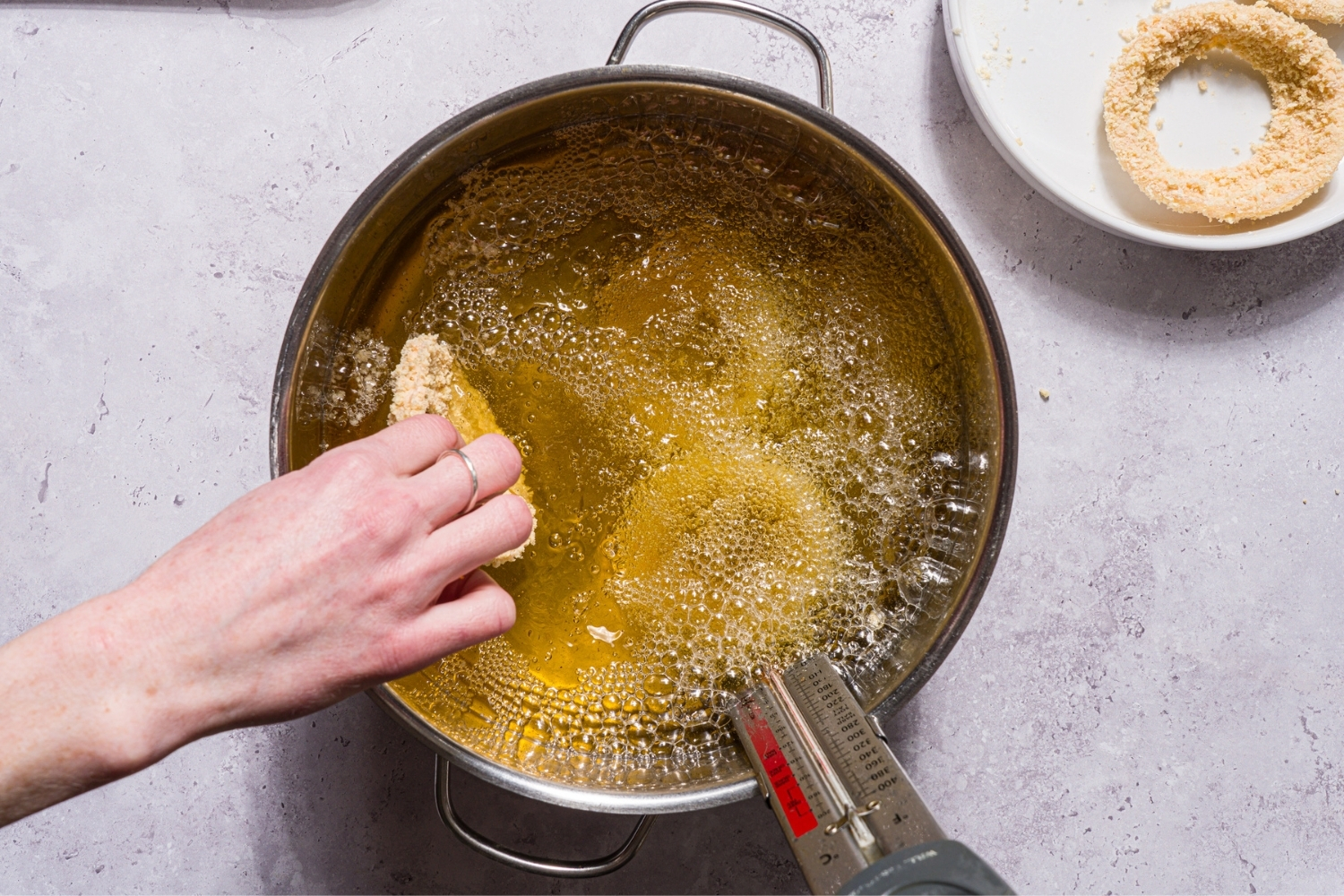 A pot with breaded onion rings frying in oil. A hand is placing an onion ring into the pot. The pot is on a stone counter.