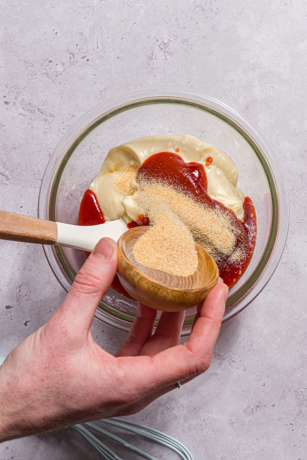 A glass bowl with ingredients to make boom boom sauce including mayo, ketchup, sriracha, and chili sauce. A hand is pouring seasonings from a small bowl into the mixture.